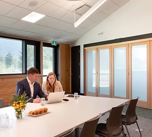 Two people working at a long table in a bright meeting room with folding glass doors at The Confluence